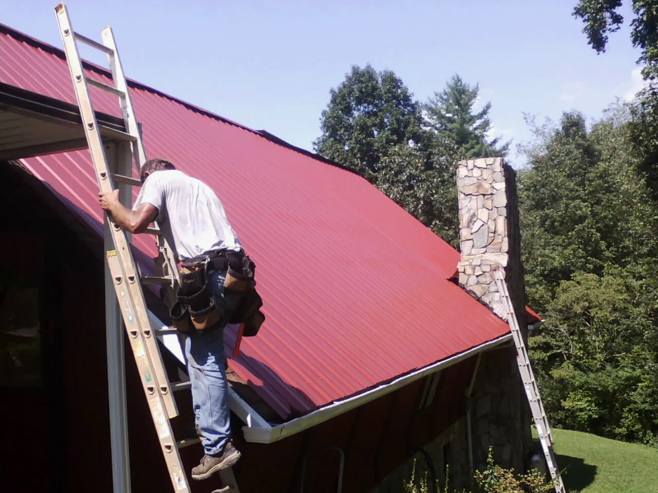 Roofer on a ladder beside a red metal roof and stone chimney — Soffit & Fascia Repair by Roofing Brothers in Hawk Point